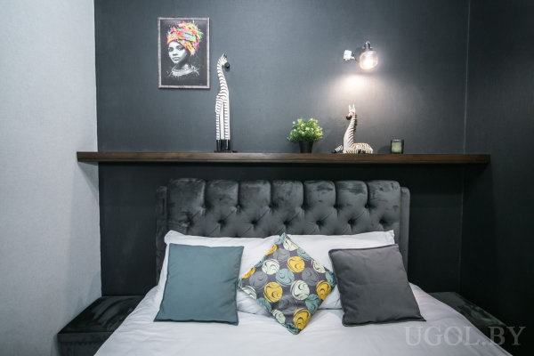 Кровать с серым изголовьем и белым бельём, декоративные подушки, деревянная полка с фигурками и растением, над ней портрет в ярких тонах, современный светильник.