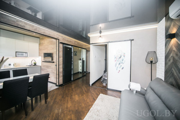 Белая кухня с встроенной техникой, обеденный стол с чёрными креслами, гостиная с серым диваном и деревянной панелью на стене, современное освещение и второй уровень с балюстрадой.