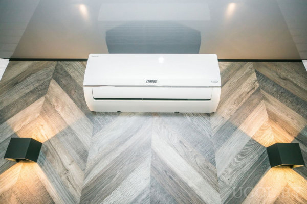 Настенный кондиционер «Zanussi» на фоне панели в ёлочку, по бокам современные светильники с тёплым светом, отражающий потолок.