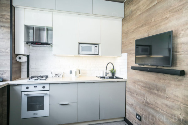 Бело-серая кухня с плитой и вытяжкой, мраморный фартук, встроенная микроволновка, чёрная мойка с растением рядом, телевизор и саундбар на деревянной панели.