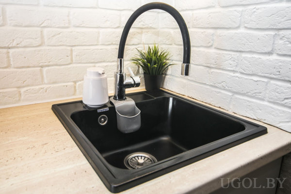 Чёрная квадратная мойка с высоким смесителем, фильтр для воды, контейнер для губки, растение и фон из белого кирпича.