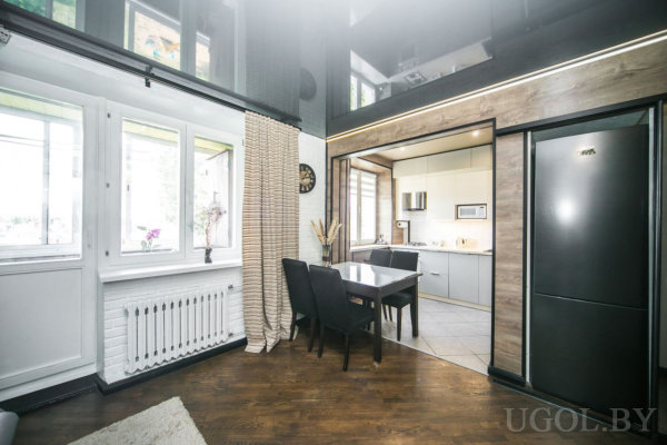 Обеденный стол с чёрными креслами, кухня с серыми шкафами и встроенной техникой, большой чёрный холодильник, белая кирпичная стена с окнами и радиатором.