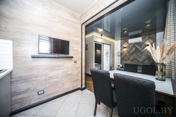 Стеклянный стол с чёрными креслами и вазой с сухоцветами, телевизор на деревянной панели, за перегородкой комната с деревянной стеной и кондиционером.