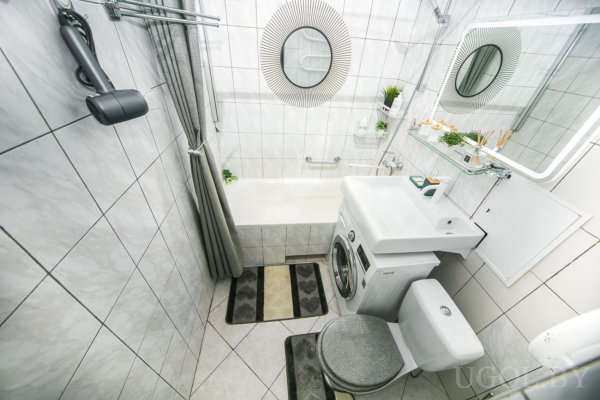 Ванна с шторой, стиральная машина рядом, унитаз и маленькая раковина над машиной, зеркало с рамкой и полки с декором.