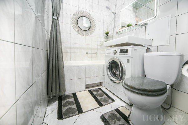 Ванна с серой шторой, стиральная машина под раковиной, унитаз рядом, зеркало с полкой и растениями, три коврика в чёрно-белой гамме.