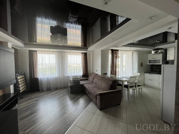 Открытая планировка с коричневым диваном, белой столовой зоной и компактной кухней в белых тонах.