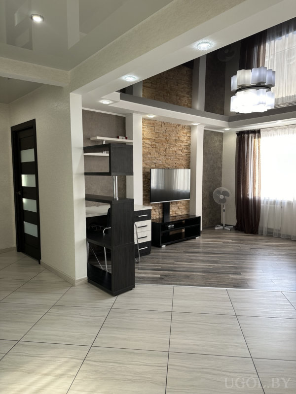 Телевизор на фоне каменной стены, рабочий стол слева, глянцевый потолок и светлая плитка.