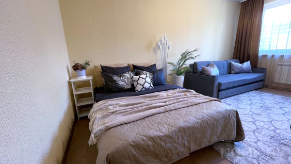 Кровать с подушками, белая тумбочка с растением, декоративные ветки и серый диван у окна.