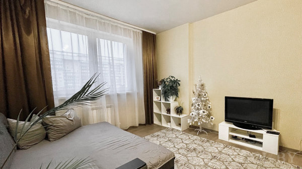 Серый диван, белая ёлка с шарами, ТВ на белой тумбе, окно с тюлем и коричневыми шторами.