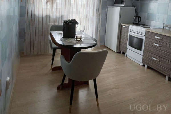 Холодильник, плита и стол на двоих на фоне светло-голубой плитки и окна с полосатыми тюлями.