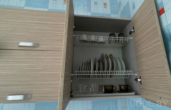 Открытый шкаф с двумя металлическими сушилками, где аккуратно размещены чашки, миски и тарелки на фоне плитки с цветными вставками.