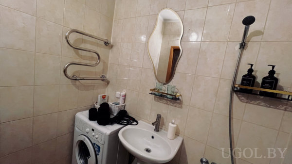 Компактное помещение с раковиной, зеркалом необычной формы, душем и настенным полотенцесушителем над стиральной машиной.