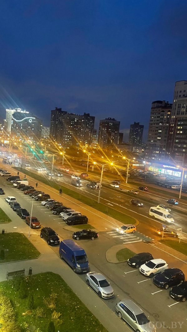 Оживлённая многополосная дорога и парковка, высотные дома с подсветкой и вывеска «SANFORBELLE» на фоне тёмного неба.