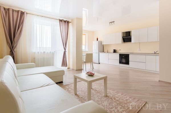 Современная гостиная‑кухня с белым угловым диваном, стеклянным столиком, светлым ковром и кухонной зоной с белыми шкафами.