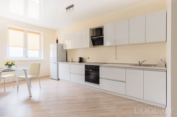 Светлая кухня с белыми шкафами, плитой, холодильником, стеклянным столом и окнами с оранжевыми жалюзи.