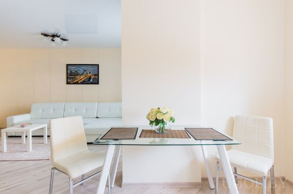 Комната с белым диваном, стеклянным столом с цветами, картиной ночного города и светлым деревянным полом.