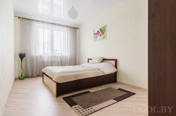 Минималистичная спальня с одинарной кроватью, ковром с узором, вазой с фиолетовыми ветками и картиной с цветами.