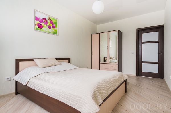 Светлая спальня с двуспальной кроватью, бежевым покрывалом, картиной с розовыми цветами и шкафом с зеркальными дверцами.