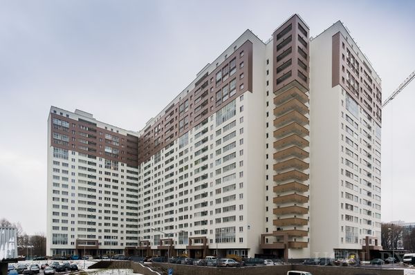 Современный жилой комплекс с белыми и коричневыми фасадами, множеством окон и балконов, парковкой и деревьями.