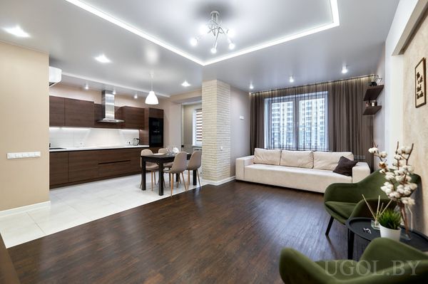 Открытая гостиная с бежевым диваном, двумя зелёными креслами, круглым столом и кухней с тёмными фасадами.