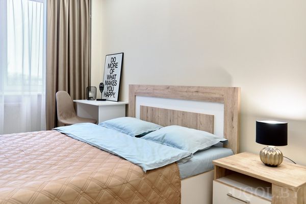 Светлая спальня с голубым текстилем, деревянной тумбочкой, белым столом и креслом у окна с бежевыми шторами.