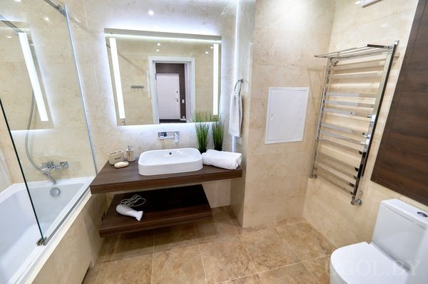 Ванная с мраморной плиткой, белой раковиной на деревянной тумбе, зеркалом с подсветкой и полотенцесушителем.