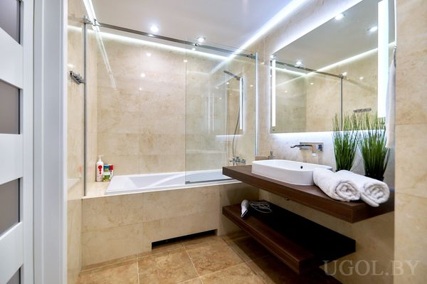 Современная ванная с ванной за стеклом, белой раковиной, декором на тумбе и подсветкой вокруг зеркала.