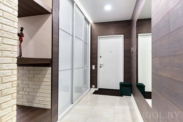 Прихожая с белой дверью, зеркальным шкафом, тёмной отделкой и скамьёй на фоне кирпичной ниши с полками.