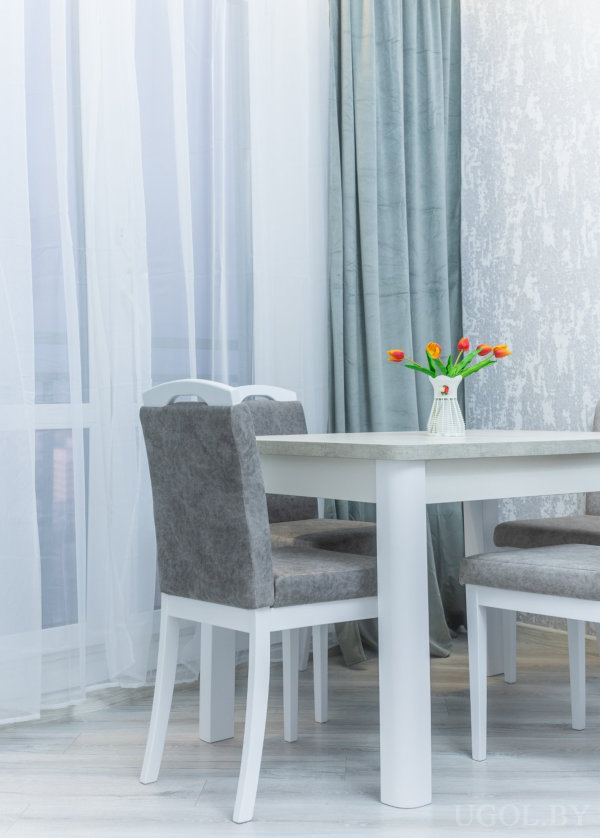 Белый стол с серыми стульями и вазой с оранжево-жёлтыми тюльпанами на фоне серых штор и светлого пола.