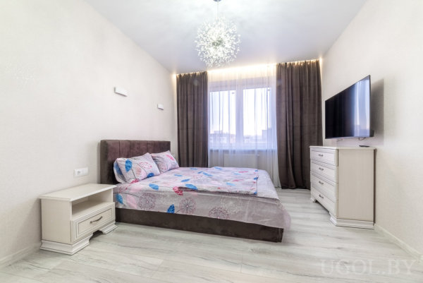 Кровать с узорным постельным бельём, белая тумбочка и телевизор на фоне окна с коричневыми шторами.