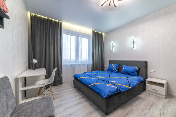 Кровать с синим покрывалом, белый стол с лампой и серое кресло на фоне штор с LED-подсветкой.