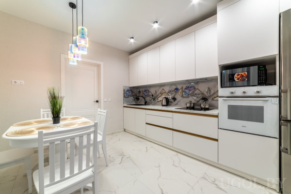 Белый гарнитур, мраморный пол, встроенная техника и стол с цветными светильниками над ним.