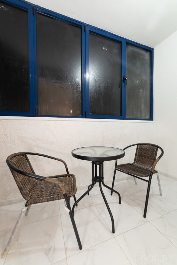 Два коричневых кресла и стеклянный столик на фоне окон с конденсатом и белых стен с текстурой.