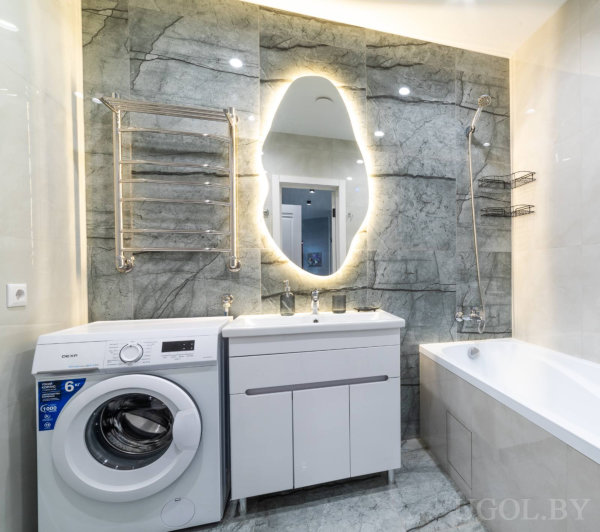 Овальное зеркало, белая тумба, ванна и серый мрамор на стенах создают современный функциональный интерьер.