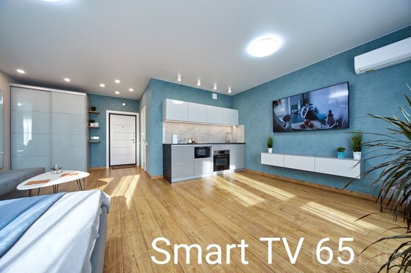 Современная гостиная с телевизором на синей стене, белой тумбой, кондиционером и кухней с белыми фасадами.