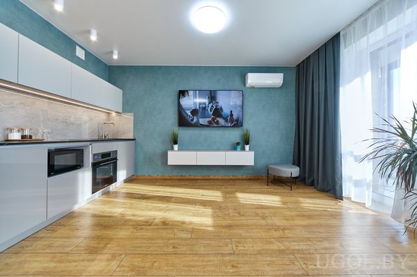 Минималистичная гостиная с телевизором на бирюзовой стене, белой тумбой, большим окном и кухней с белыми шкафами.
