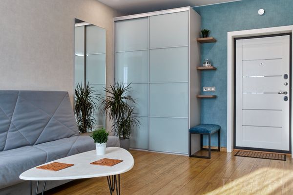 Уютная зона с серым диваном, белым столом, зеркалом, растениями и синим акцентом на стене.