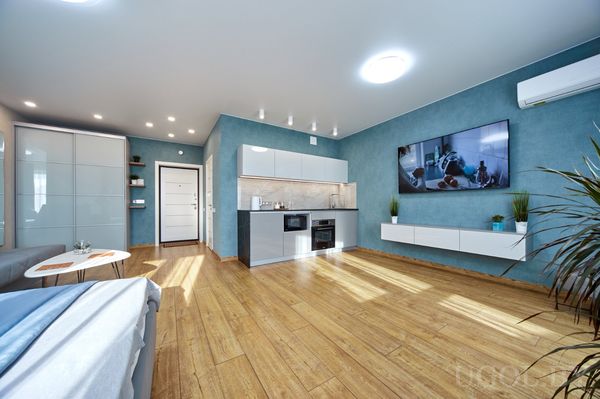 Открытая гостиная с телевизором, кирпичной колонной, белой кухней и круглым столом на фоне синей стены.