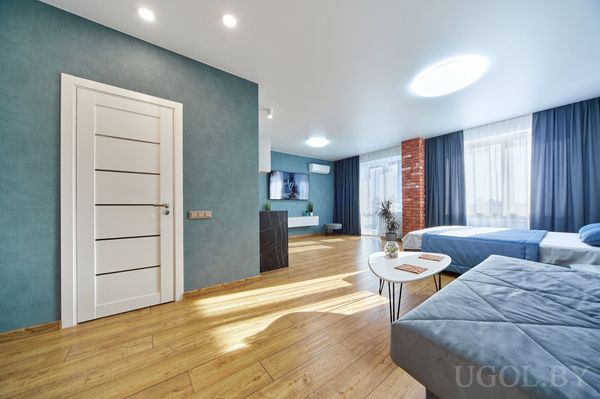 Просторная студия с кроватью, диваном, круглым столиком, кирпичной колонной и телевизором на синей стене.