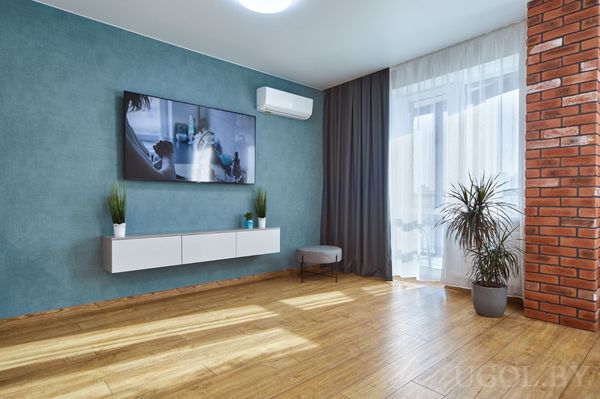 Современная гостиная с телевизором, кирпичной стеной, серым пуфом и растениями у окна с тюлем и шторами.