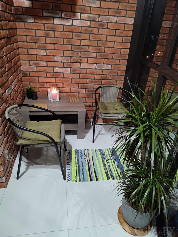 Уютная зона с двумя креслами, деревянным столом, свечами, растениями и видом на город через стекло.