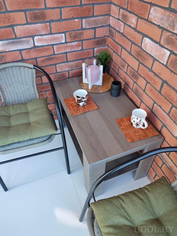 Тёплый уголок с плетёными креслами, деревянным столом, свечой, кружками и кирпичной стеной.