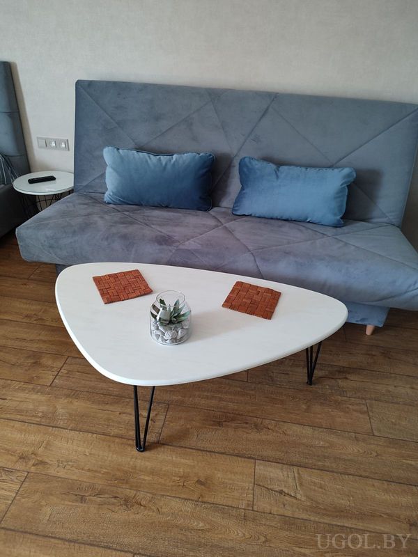 Минималистичная гостиная с серым диваном, треугольным столиком, растением и деревянным полом.