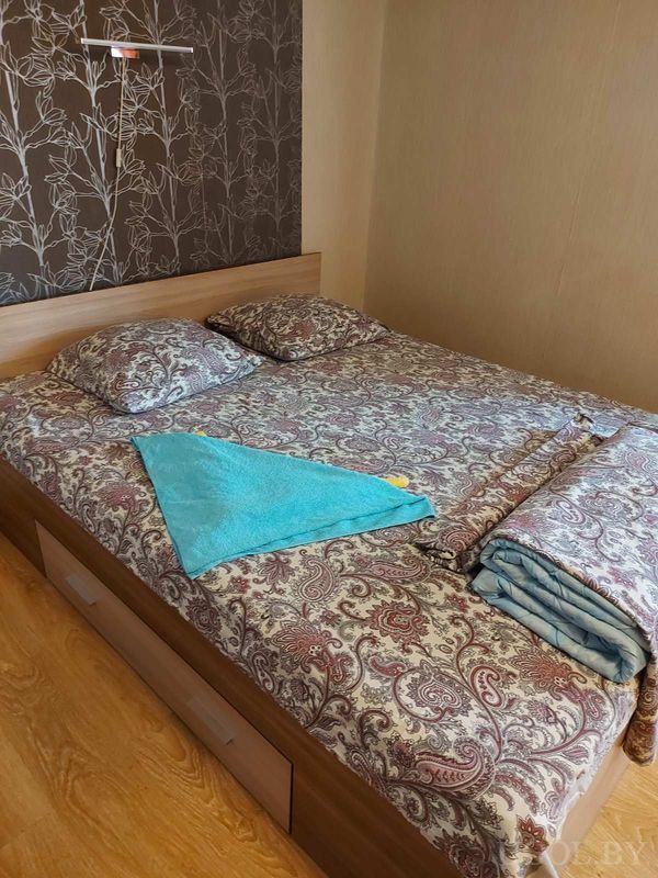 Интерьер с деревянной кроватью, тёмной стеной с панно, светильником, синим полотенцем и голубым пледом.