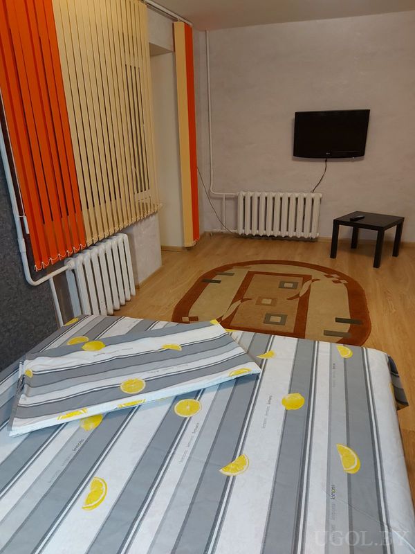 Интерьер с кроватью, текстилем с лимонами, телевизором, ковром и окнами с жалюзи в оранжево-бежевых тонах.