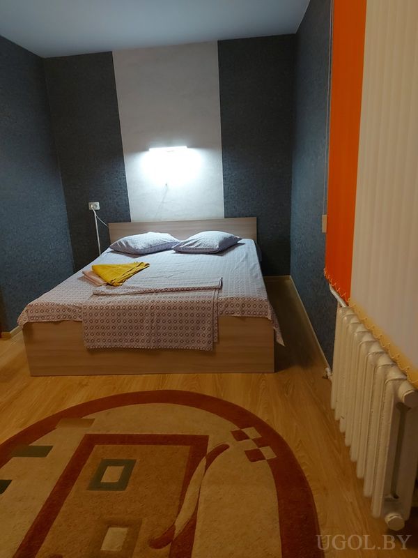 Комната с кроватью, тёмной стеной, светильником, ковром с узором и окном с вертикальными жалюзи.