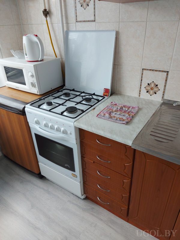 Компактная кухня с белой плитой, микроволновкой, чайником, мойкой и фартуком с цветочным рисунком.