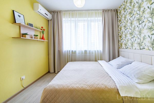 Современная спальня с бежевым покрывалом, жёлтой стеной, подвесной полкой с вазой, растениями и картинами.