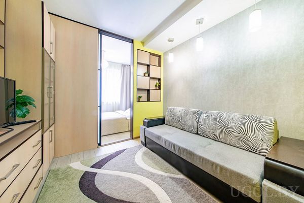 Компактная гостиная с длинным диваном, ковром с узором, полками с декором и видом на спальню через раздвижную дверь.