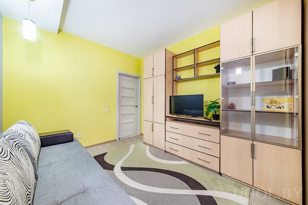 Гостиная с жёлтой стеной, серым диваном, ковром с завитками и большим деревянным шкафом с телевизором и декором.